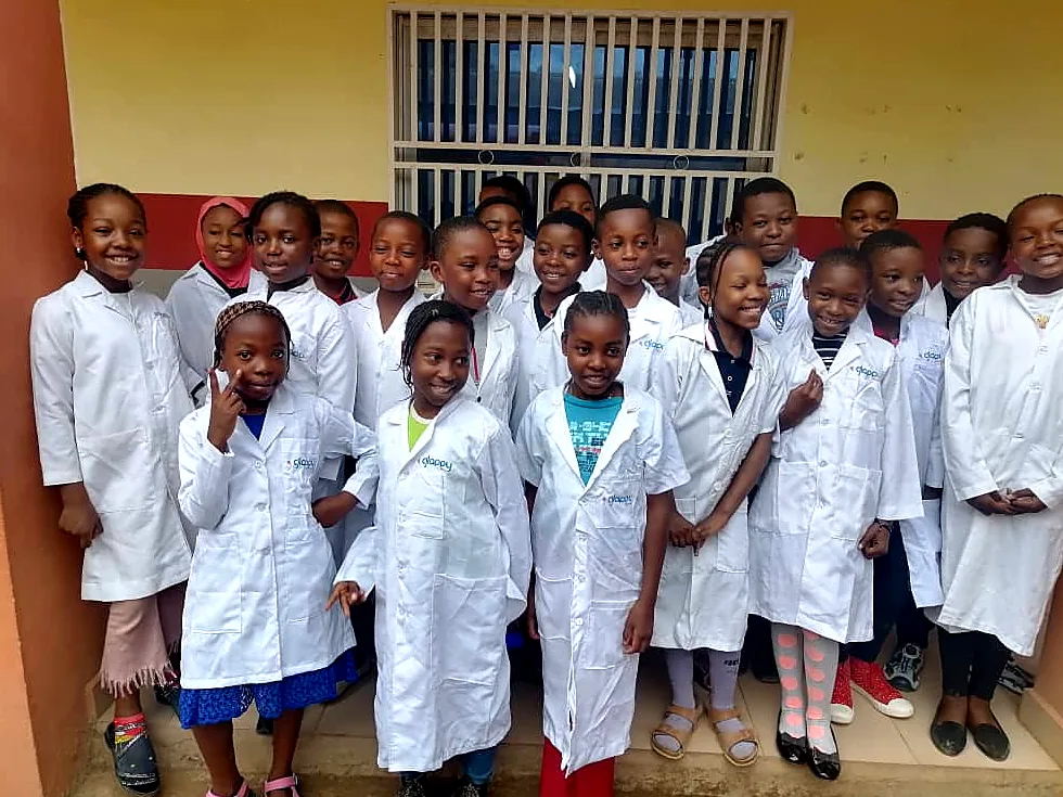 Rows of smiling children in white lab coats posing together outside their classroom