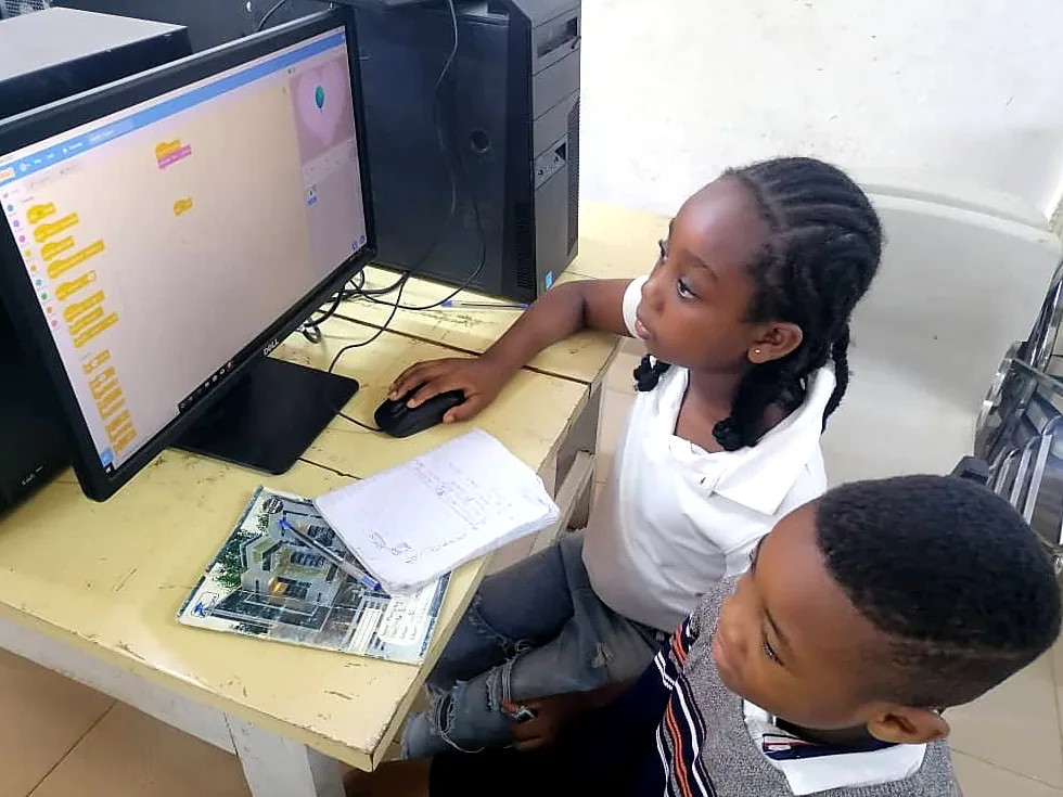 A girl works through a block coding lesson on a desktop computer while a classmate watches