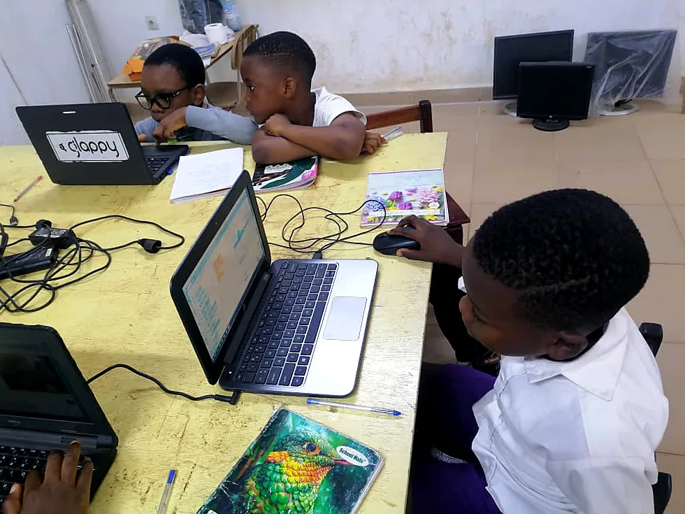 A boy studies his laptop screen at a shared desk, with other students working behind him