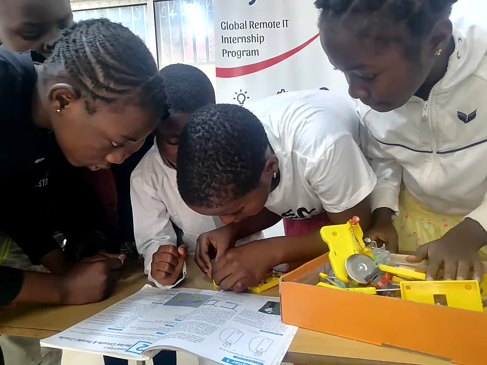 Students in white lab coats lean over a yellow robotics kit, reading the build instructions together